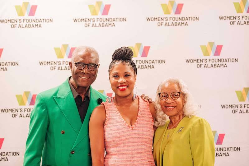 Three people pose and smile in front of a step-and-repeat banner displaying the Women's Foundation of Alabama logo, celebrating the announcement of their new CEO.