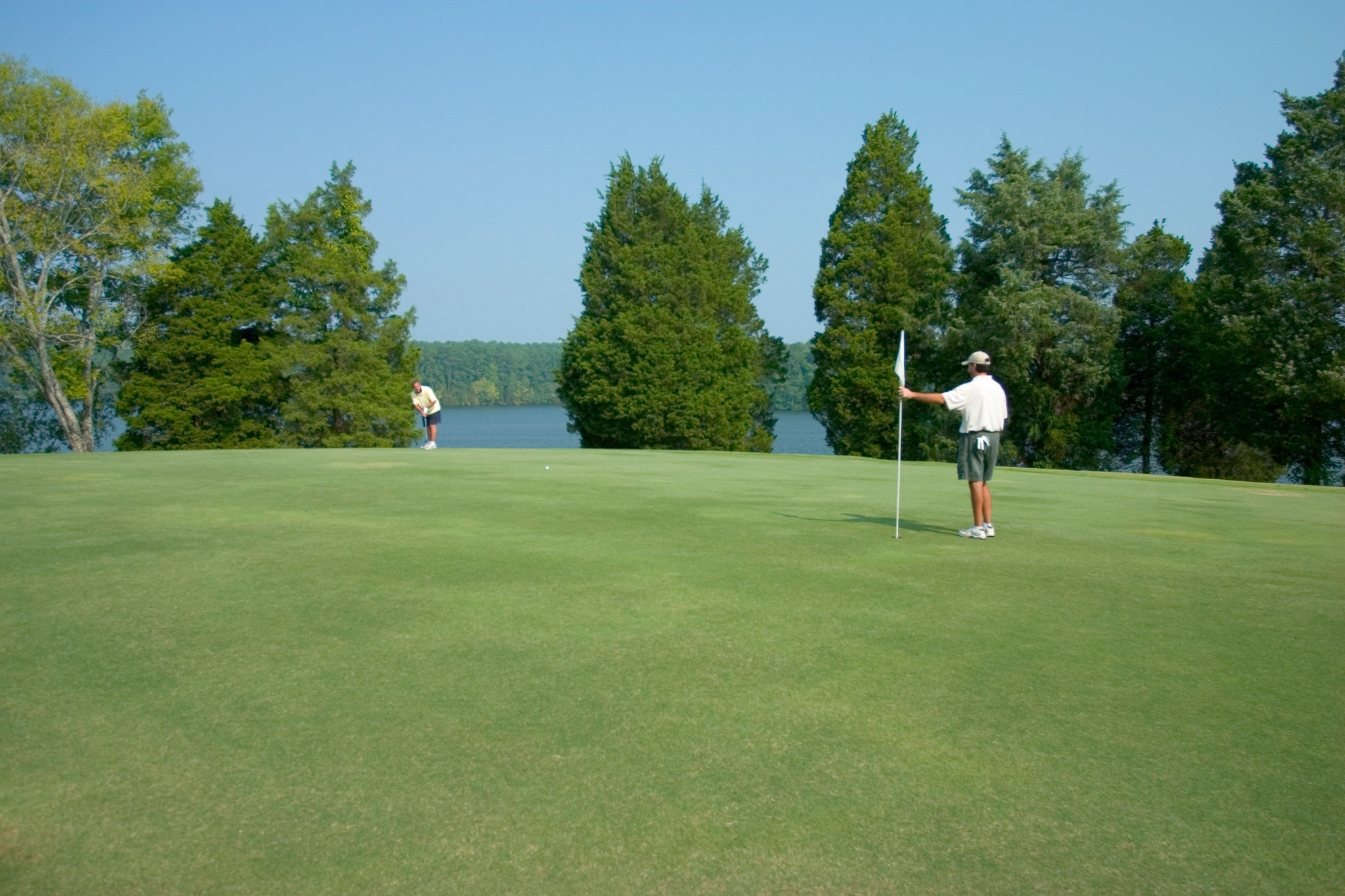 Two people are on a golf course green at The General Golf Course in Joe Wheeler State Park; one holds the flagstick while the other prepares to putt. Trees and a lake are visible in the background.