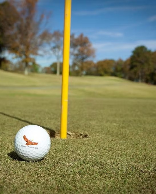 A golf ball rests on the green near the hole with the flagstick, set against a backdrop of trees and blue sky at Eagle's Nest Golf Course in Lake Guntersville State Park.