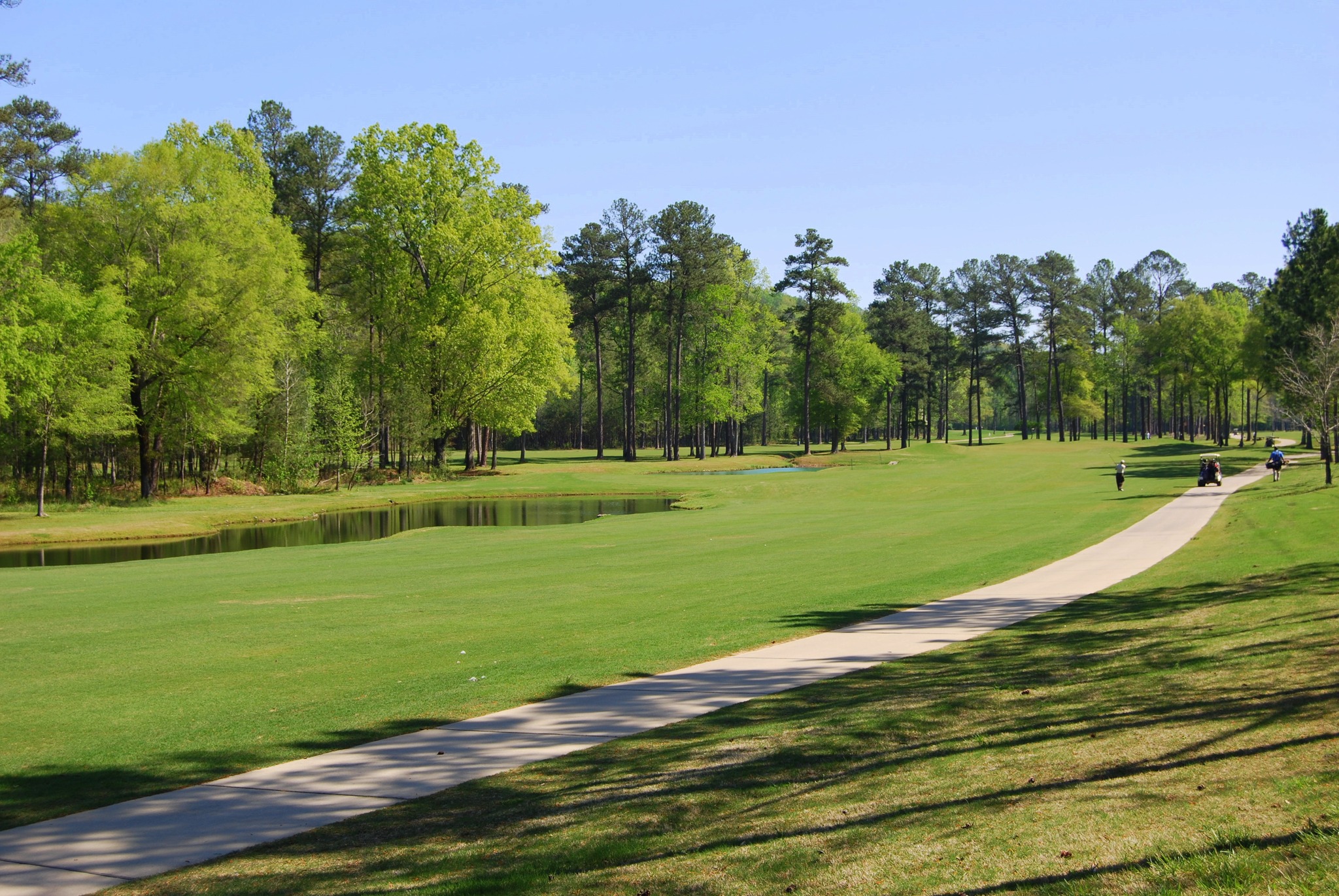 A paved path runs alongside The Oaks golf course at Oak Mountain State Park with trees and a pond under a clear blue sky.