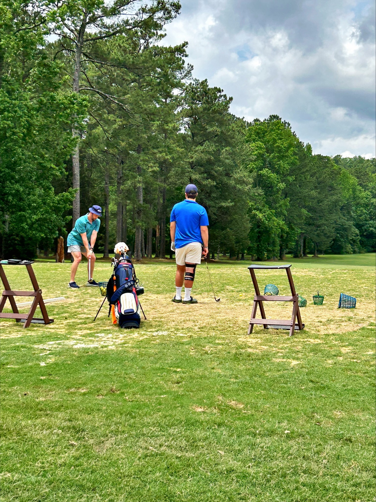 Two people on a golf driving range at The Oaks Golf Course in Oak Mountain State Park, one preparing to hit a ball while the other stands nearby; golf bags, buckets, and folding chairs sit on the grass.