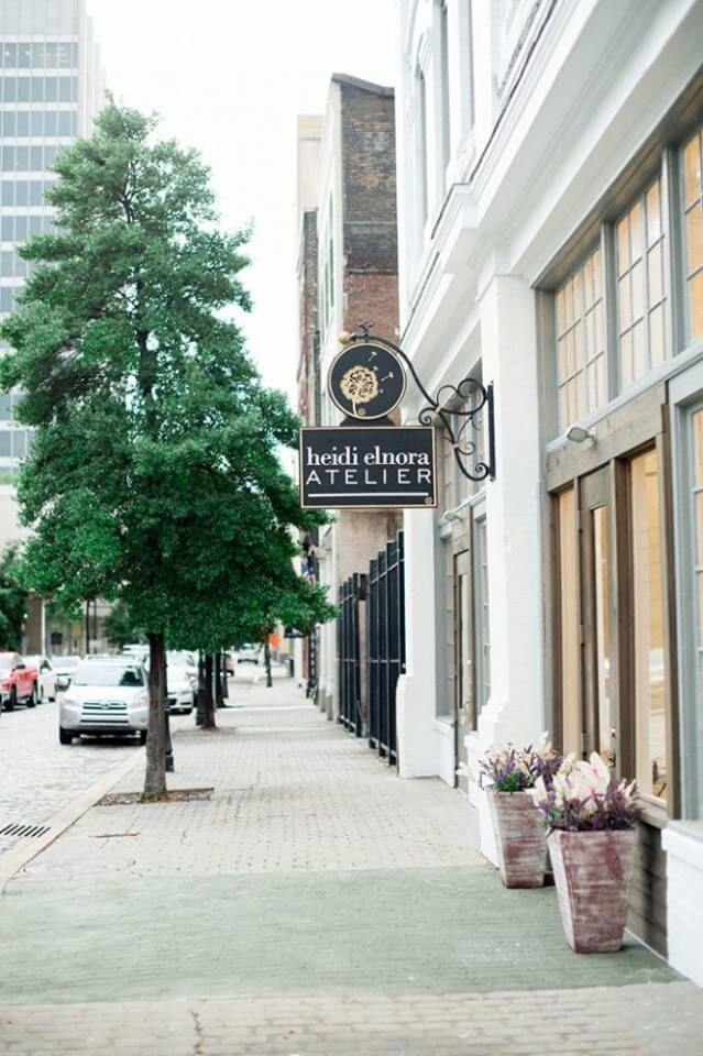 A sidewalk view of a city street features a "Heidi Elnora Atelier" sign above the entrance to a white building; potted plants are near the door.