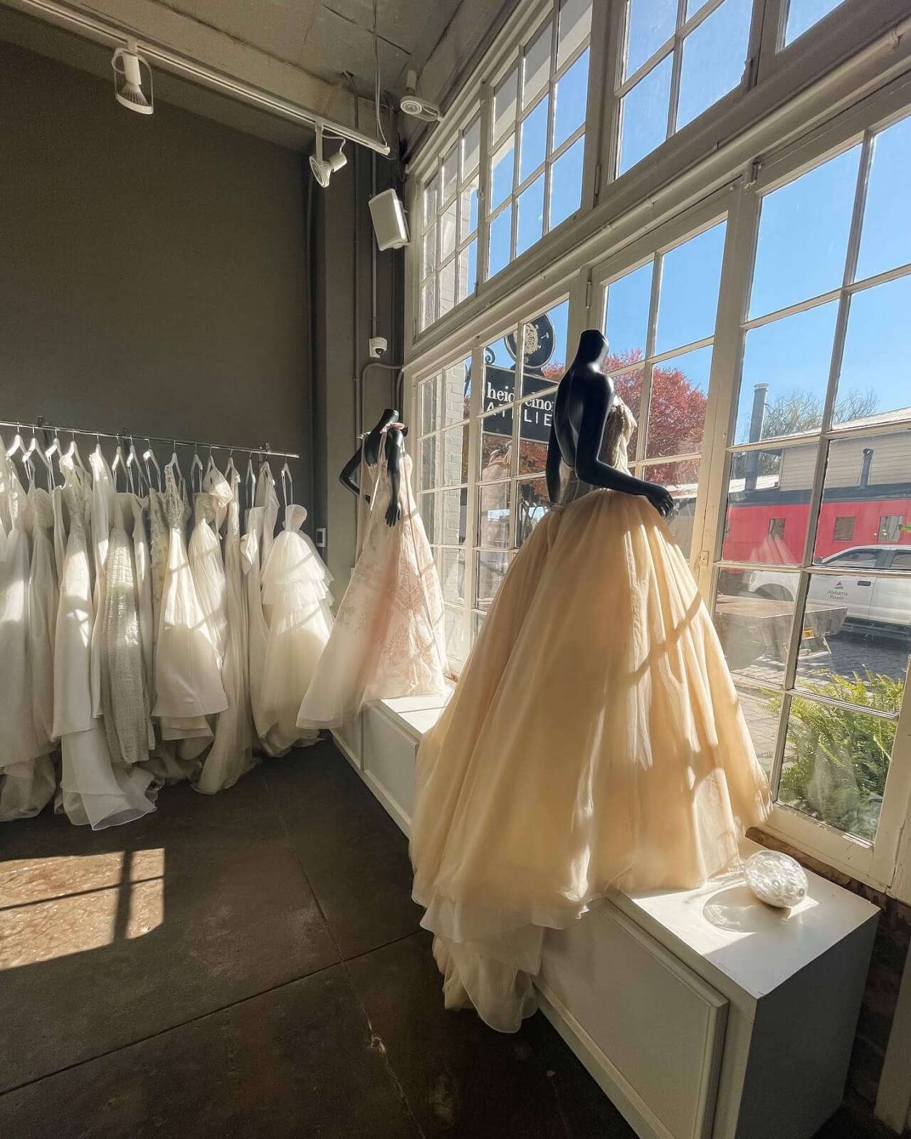 A bridal shop interior with Heidi Elnora wedding dresses on hangers and two mannequins in gowns near large atelier-style windows with sunlight streaming in.