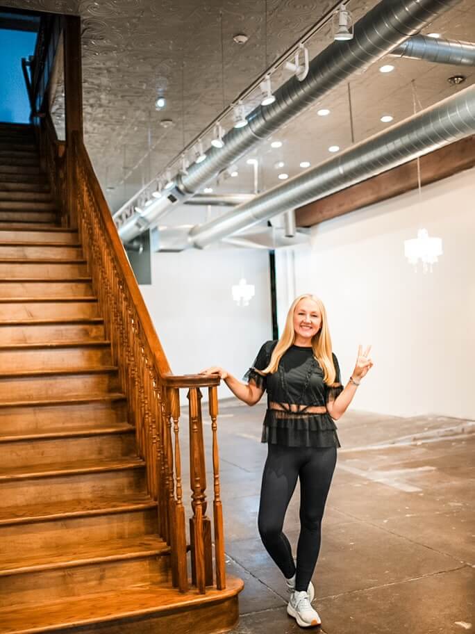 Heidi Elnora in black workout clothes stands next to a wooden staircase in her historic building on Morris Avenue which she lovingly restored.