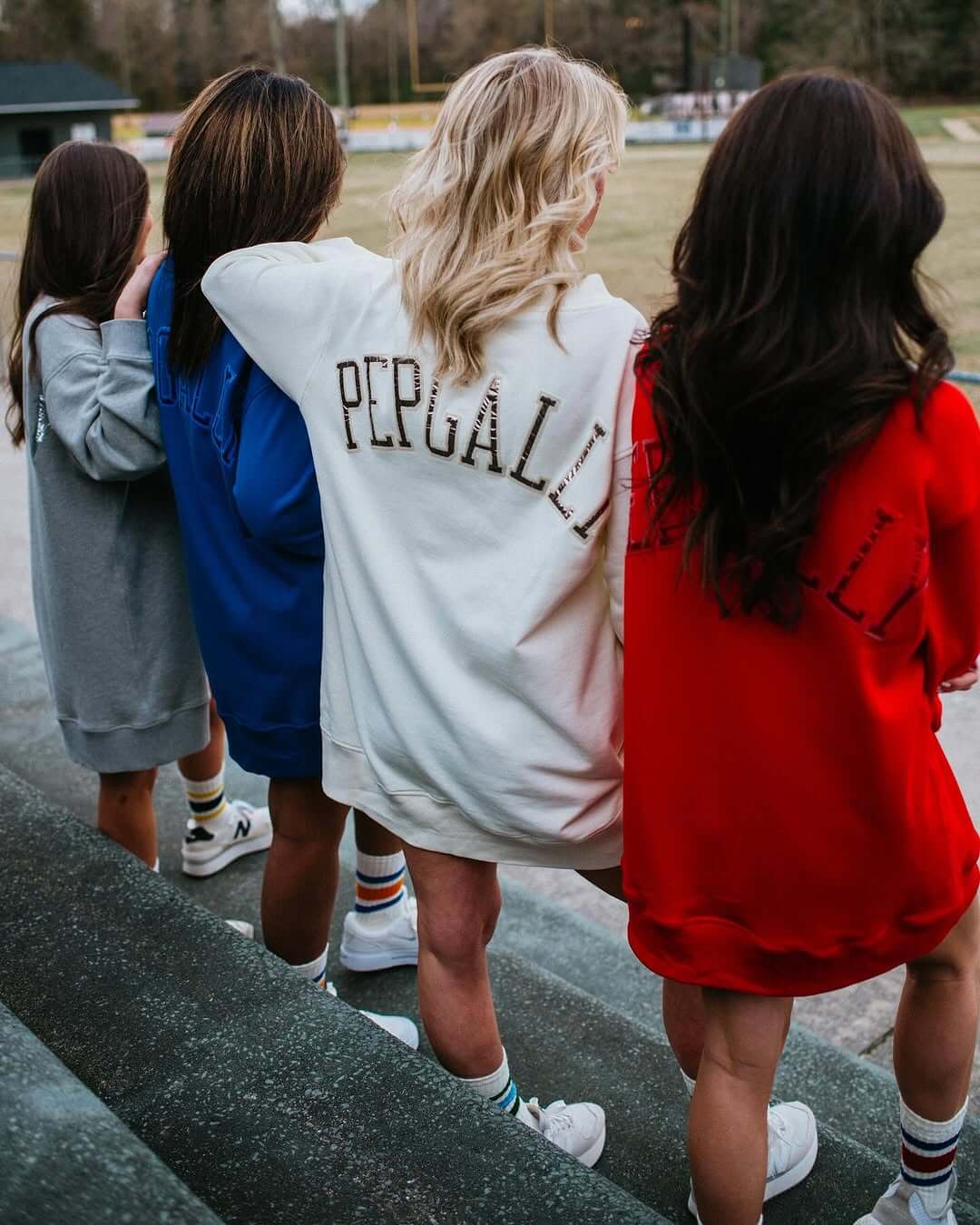 Four women stand on outdoor steps facing a sports field, each in oversized sweatshirts—gray, blue, white, and red, with "PEPGALLY" on the back. 