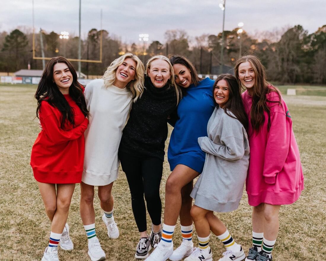 Six young women stand outdoors on a sports field, smiling and posing together. They wear oversized sweatshirts in various colors from Pep Gally and sneakers with striped socks. Heidi Elnora wears all black in the middle.
