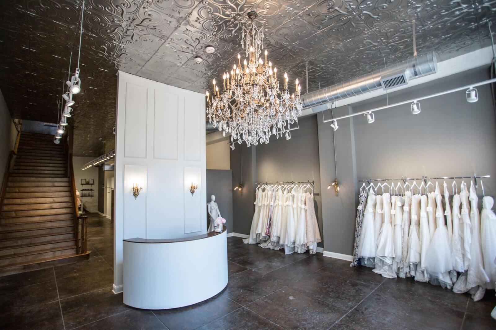 Inside Heidi Elnora Atelier with a chandelier, curved white reception desk, and racks of Heidi Elnora wedding dresses against gray walls. A staircase is visible on the left, and tin tiles line the ceiling.
