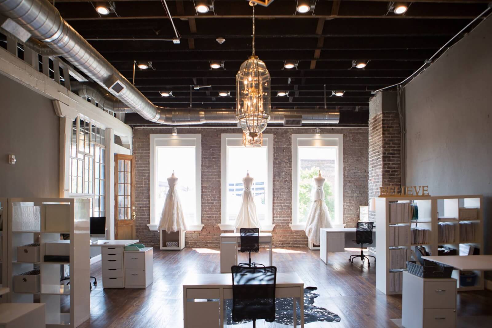 Spacious, modern bridal atelier with desks, white shelving units, and three Heidi Elnora wedding dresses displayed by large windows under a central chandelier. 