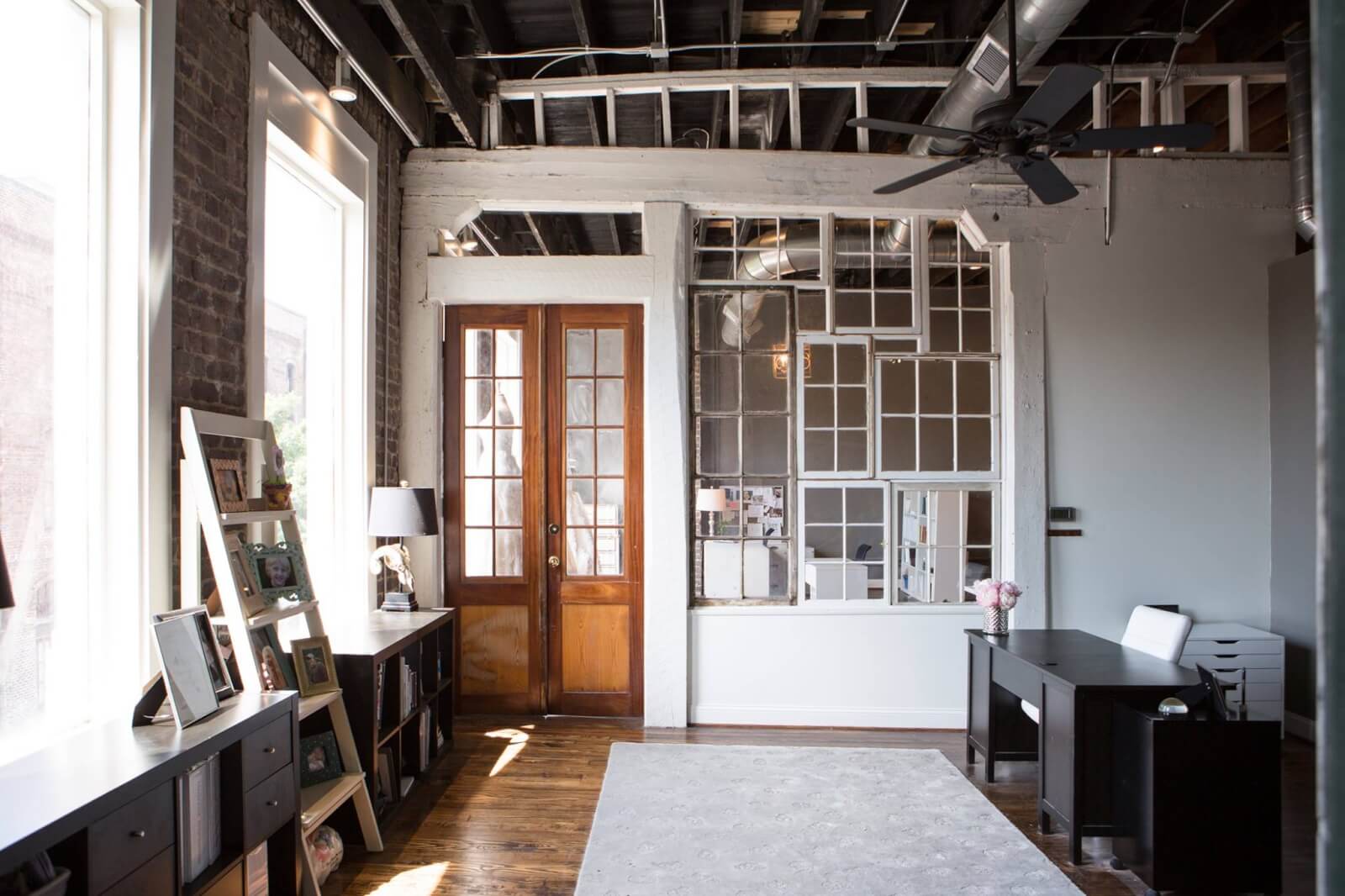 Modern office space with exposed ceiling beams, wooden floors, large windows, a desk, shelves, and a wall made of repurposed window frames.