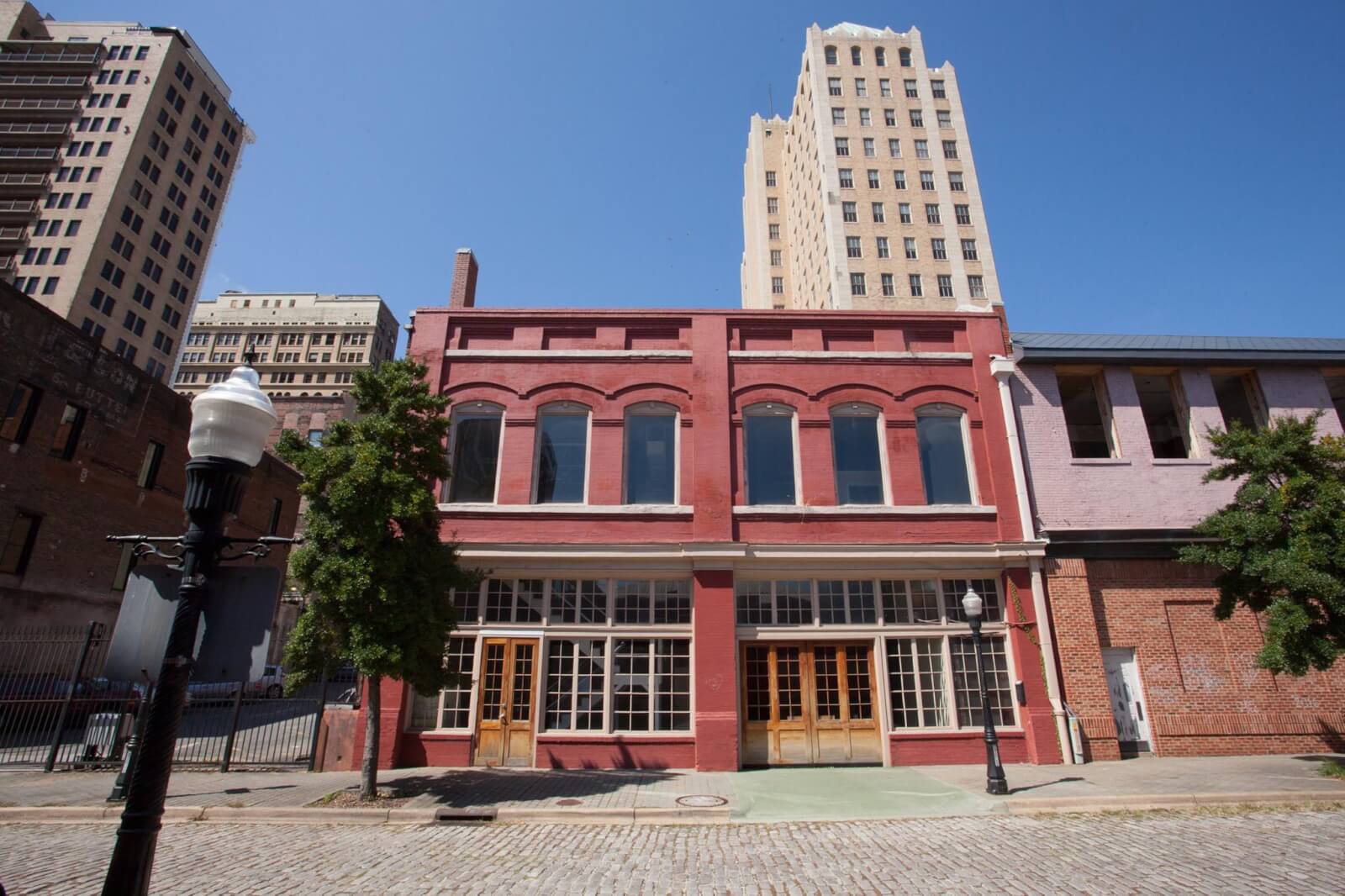 A two-story red brick building with large windows that was to become home to the Heidi Elnora Atelier, stands on a cobblestone street amid taller city buildings under a clear blue sky. 