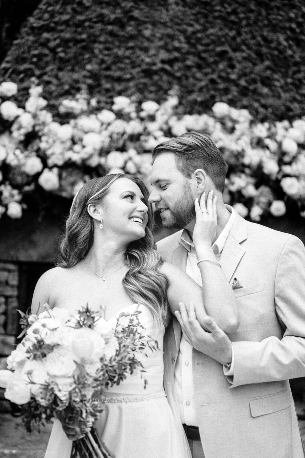 A bride holding a bouquet smiles at her groom as he gently touches her face; both are dressed formally, standing in front of a floral arrangement at their beautiful outdoor wedding.