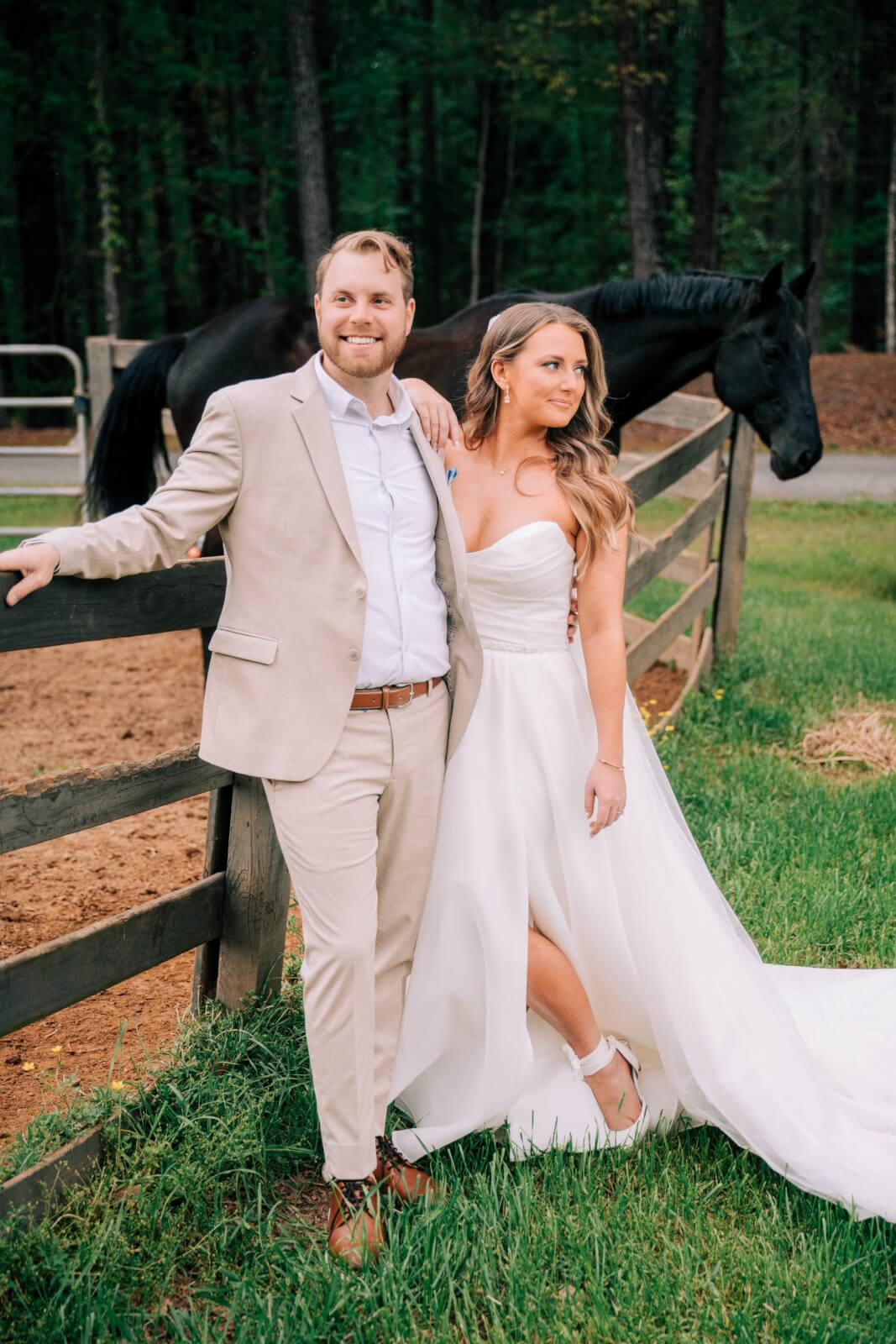 A couple in formal wedding attire stands by a wooden fence in front of a black horse, surrounded by trees, capturing the charm of an outdoor wedding.