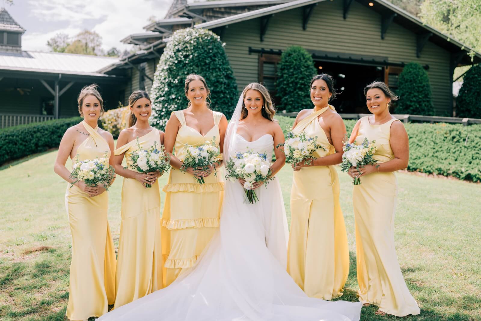Six women stand outdoors at a BHAM wedding in front of a building, all wearing yellow dresses except the bride in white. Each holds a bouquet of flowers and smiles brightly at the camera, capturing this joyful outdoor wedding moment.