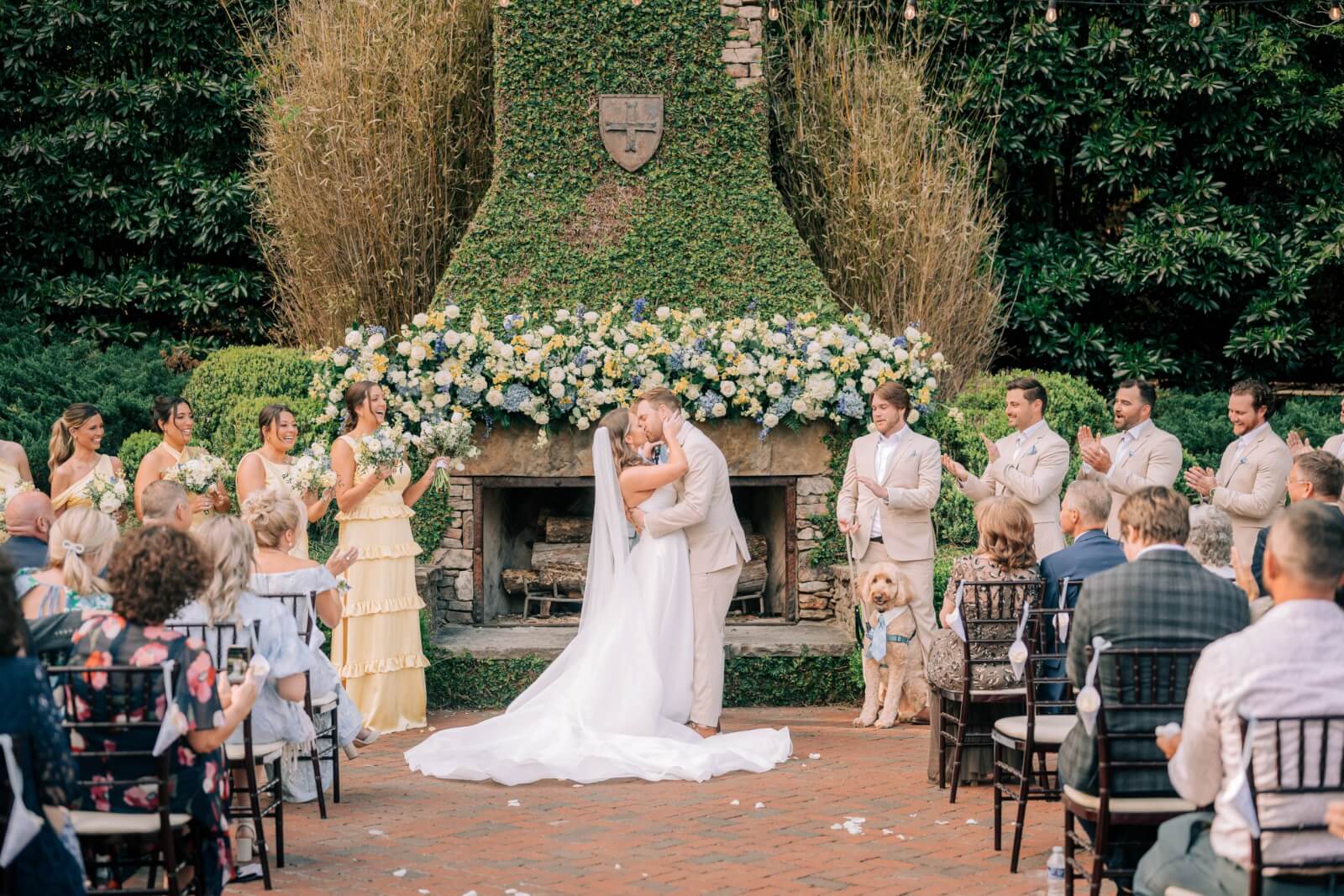 A bride and groom share a kiss at their BHAM wedding ceremony, surrounded by guests, their wedding party, and precious pups, in front of a flower-adorned stone fireplace.