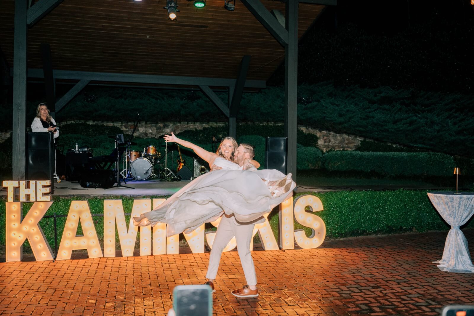 At an outdoor wedding, a bride and groom perform a dance lift at night in front of large illuminated letters spelling "THE KAMINSKIS," with a band setup visible in the background.