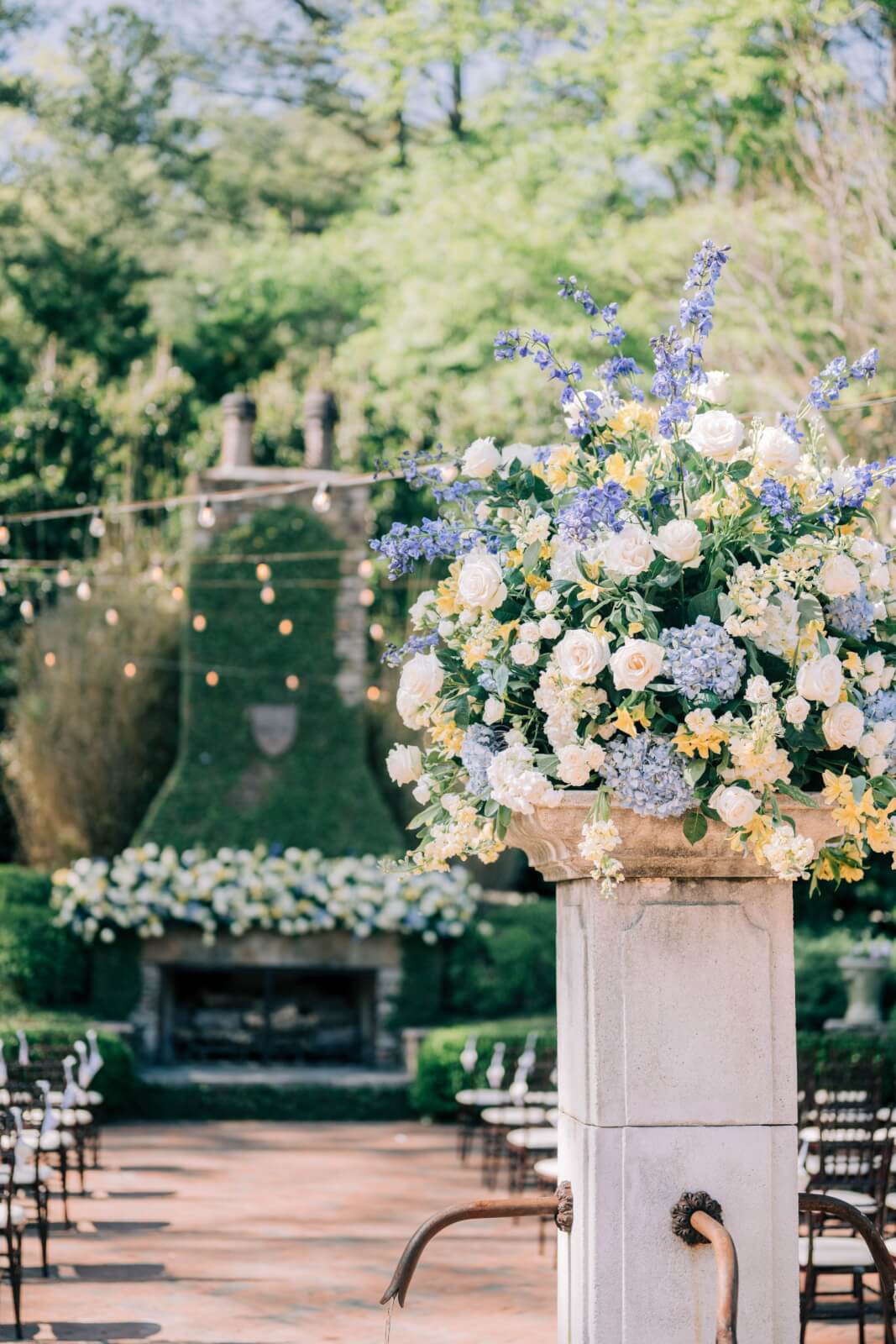 A large floral arrangement with white, yellow, and blue flowers sits on a stone pedestal at an outdoor wedding venue with string lights, rows of chairs, and all the joy of a beautiful BHAM wedding celebration.