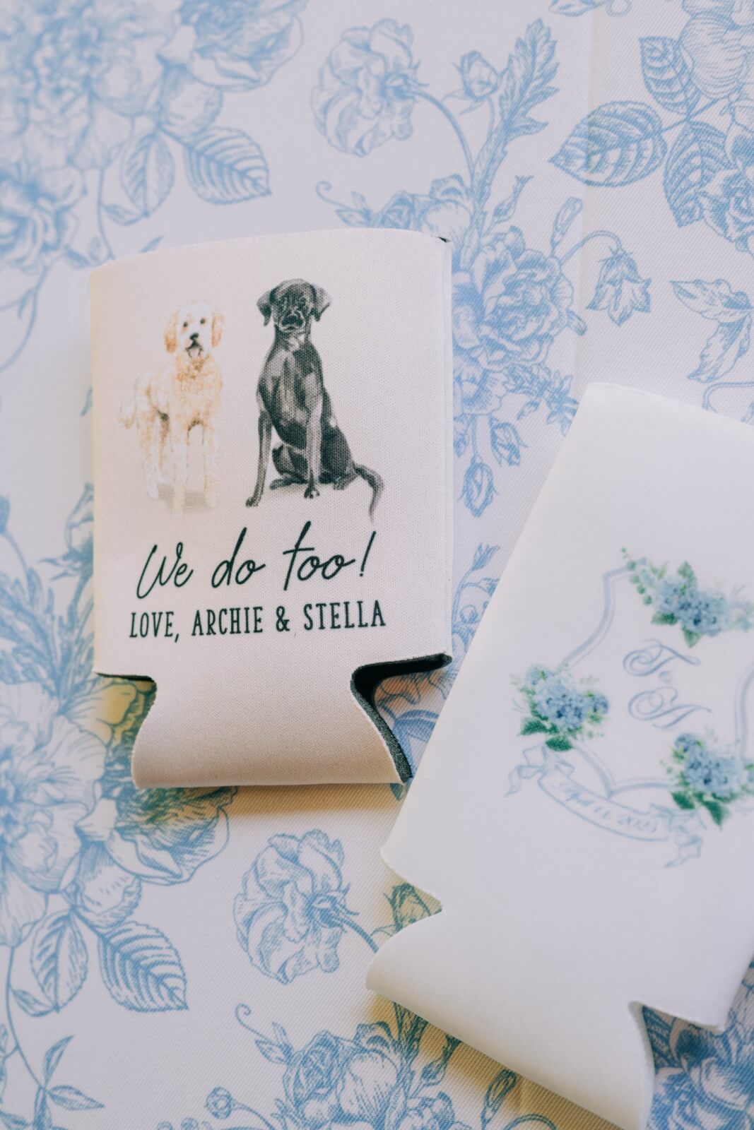 Two can coolers on a floral-patterned surface; one features illustrations of precious pups and the words "We do too! Love, Archie & Stella"—a charming detail for any outdoor wedding.