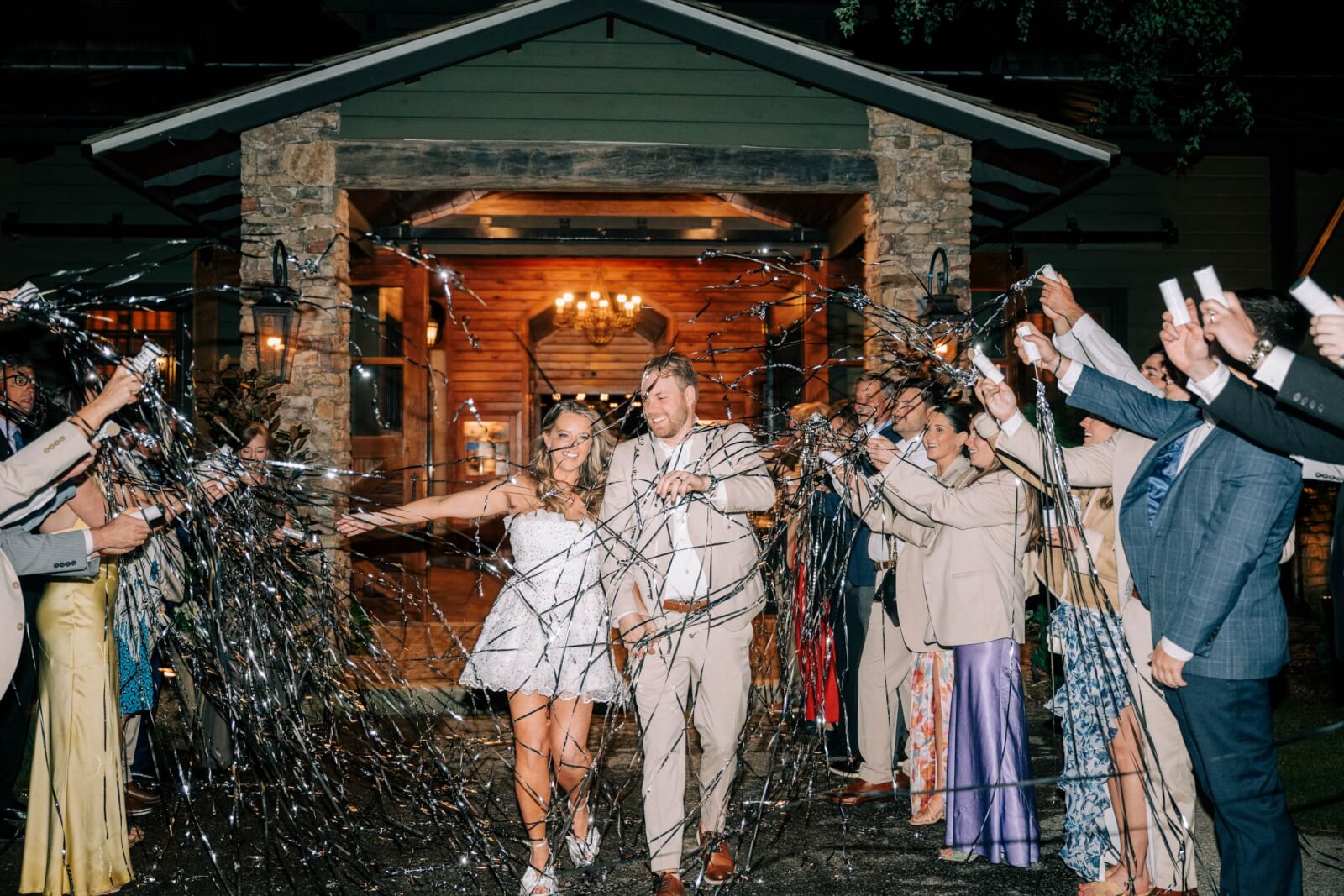 A couple in formal attire walks through a crowd of people holding streamers outside a building at night, celebrating their outdoor BHAM wedding.