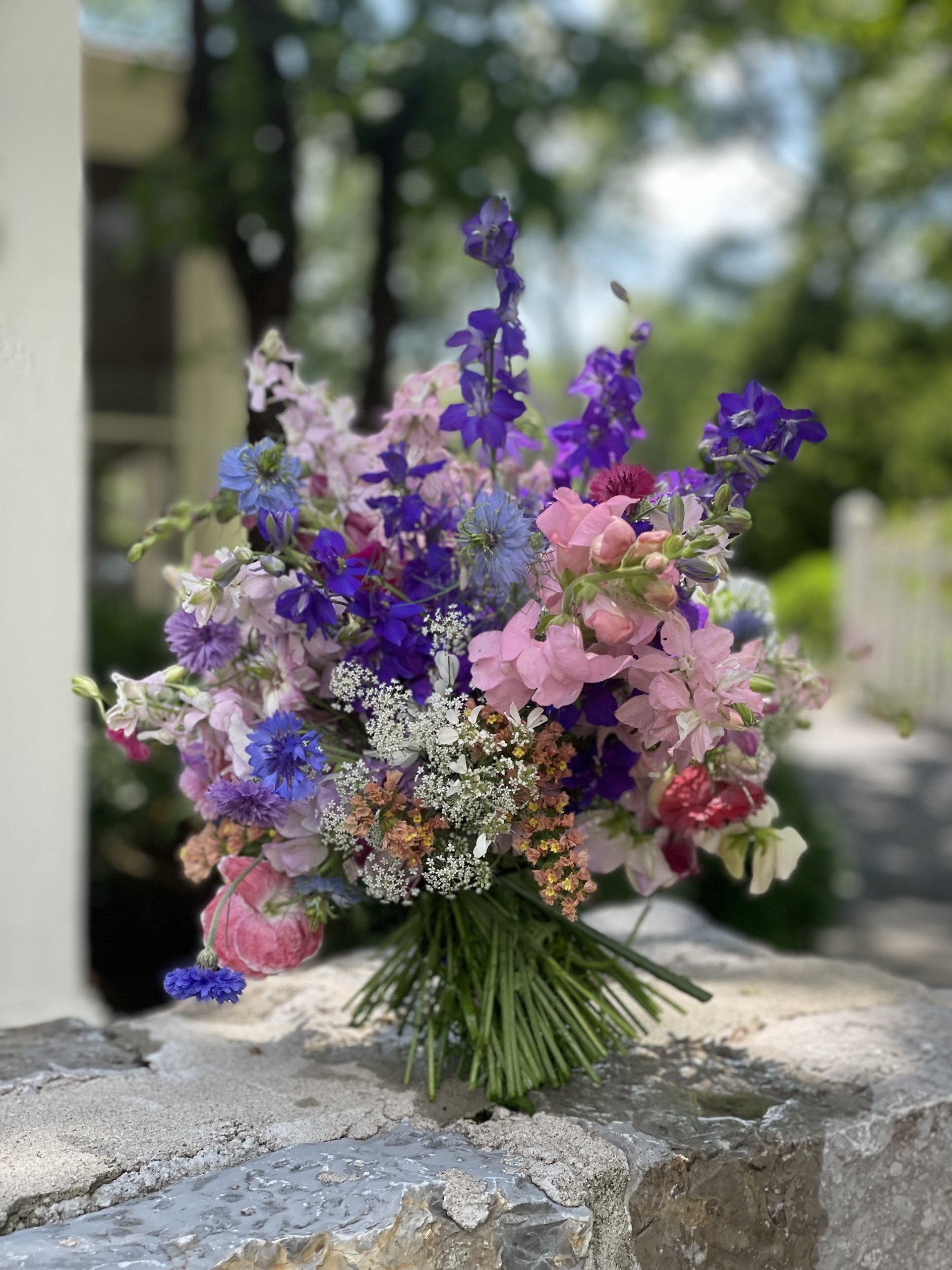 A bouquet of assorted wildflowers in shades of purple, pink, and white rests on a stone surface outdoors—a lovely display inspired by a gorgeous garden with trees and a fence in the background.