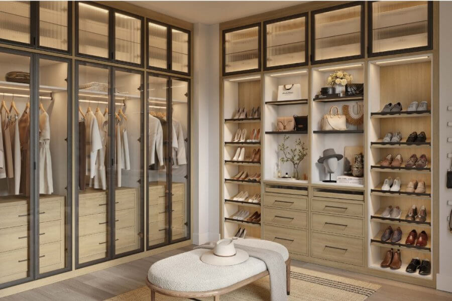 A modern walk-in closet by California Closets features glass-front cabinets, hanging clothes, shelves of shoes and accessories, wooden drawers, and a cushioned bench in the center—perfect inspiration for the upcoming Anniversary Sales Event.