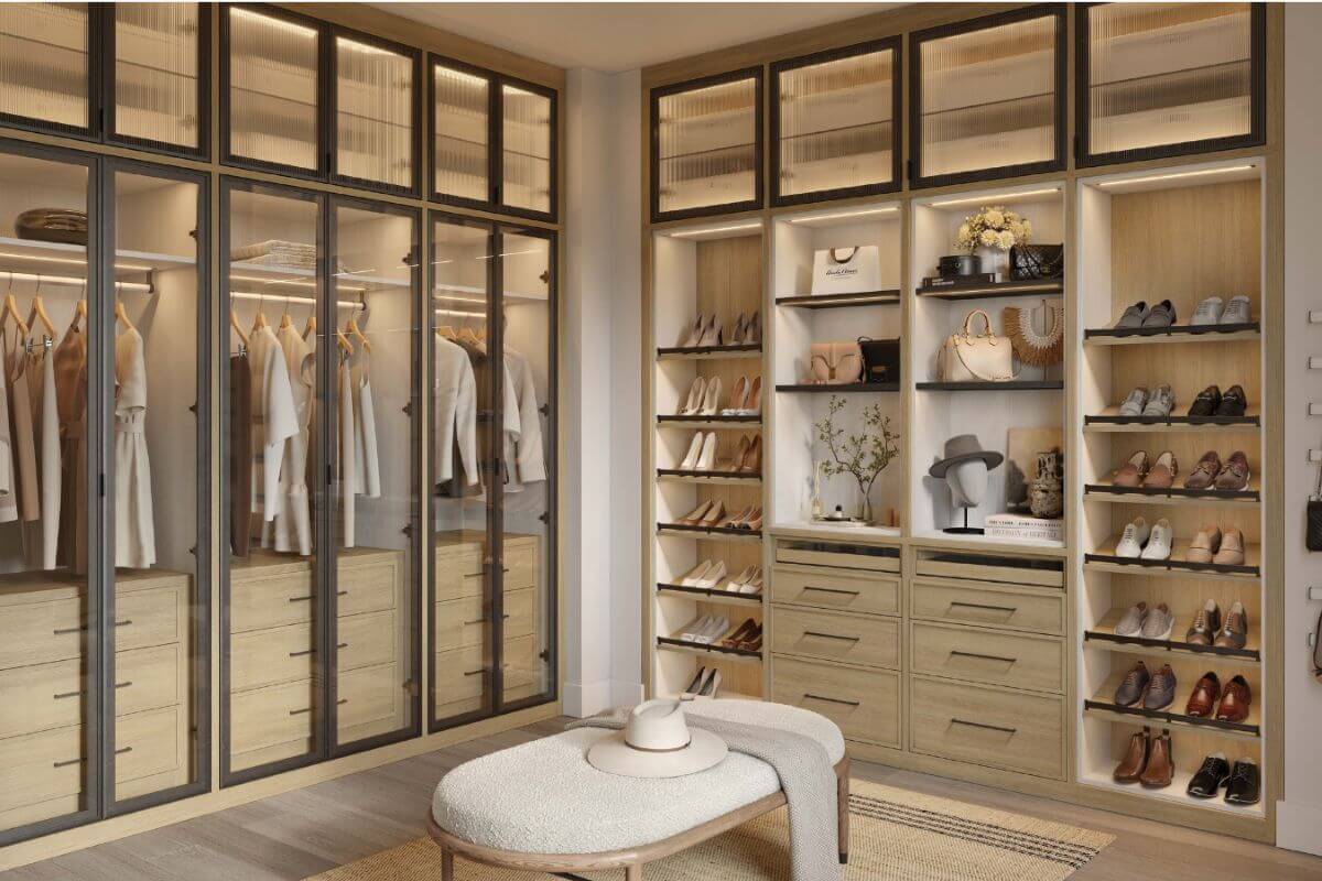 A modern walk-in closet by California Closets features glass-front cabinets, hanging clothes, shelves of shoes and accessories, wooden drawers, and a cushioned bench in the center—perfect inspiration for the upcoming Anniversary Sales Event.