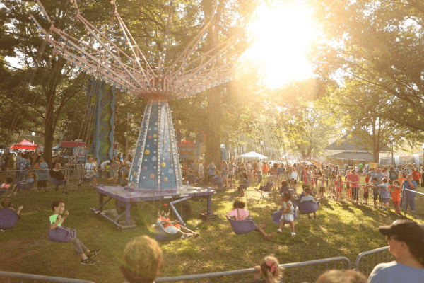 Children and adults ride a spinning swing ride at an outdoor fair during the Tennessee Soybean Festival, surrounded by trees and a lively crowd, with sunlight streaming through the branches.