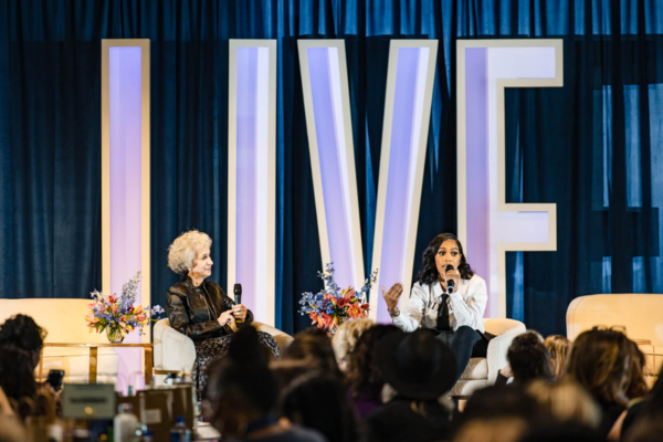 Two women sit on stage with microphones, speaking to an audience at BrainTrust LIVE. Large letters spelling "LIVE" are displayed in the background, while flower arrangements fuel the vibrant atmosphere beside each speaker.