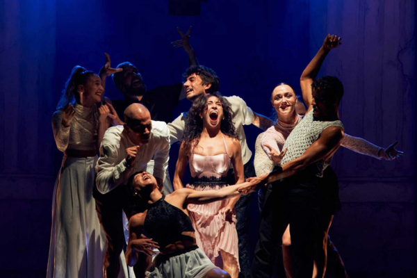 A group of eight performers from Compagnie Dyptik pose dramatically under blue lighting in Le Grand Bal, with one woman in the center appearing to sing or shout.