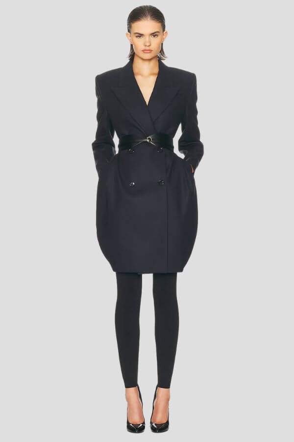 Fall 2025 Fashion: A Stylist's Lookbook of This Season's Trends - 10 A woman stands facing forward wearing a black belted coat, black leggings, and black high-heeled shoes against a plain light gray background, showcasing sleek style inspired by Fall 2025 fashion trends.