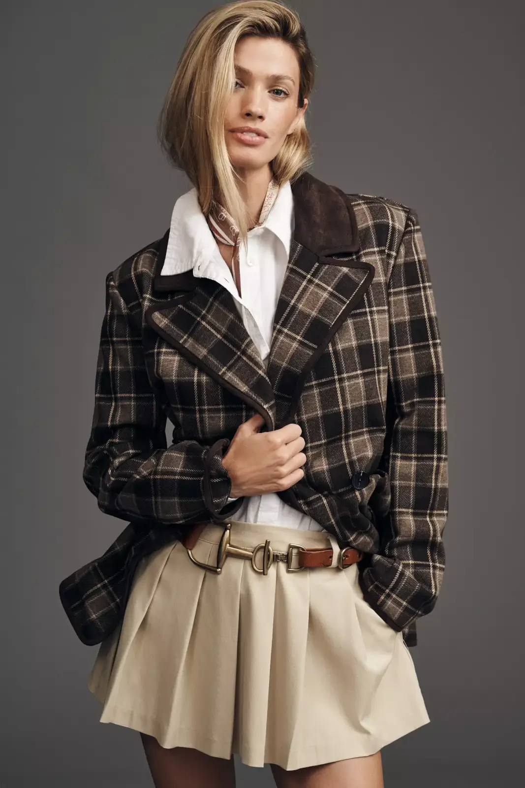 Fall 2025 Fashion: A Stylist's Lookbook of This Season's Trends - 75 A woman poses in a plaid blazer over a white collared shirt, beige pleated skirt, and brown belt against a plain gray background, capturing the timeless elegance of fall 2025 fashion.