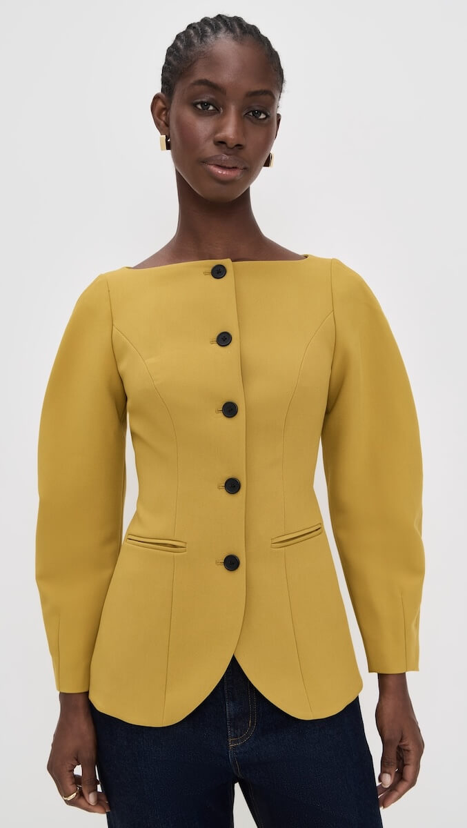 Fall 2025 Fashion: A Stylist's Lookbook of This Season's Trends - 14 A person wearing a mustard yellow, tailored jacket with black buttons and long sleeves, paired with dark jeans, stands against a plain light background, showcasing a chic look inspired by fall 2025 fashion.