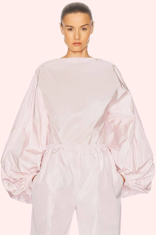 Fall 2025 Fashion: A Stylist's Lookbook of This Season's Trends - 1 A woman wearing a shiny, light pink outfit with long, voluminous sleeves and an elastic waistband—echoing Fall 2025 fashion trends—stands with her hands in her pockets against a plain background.