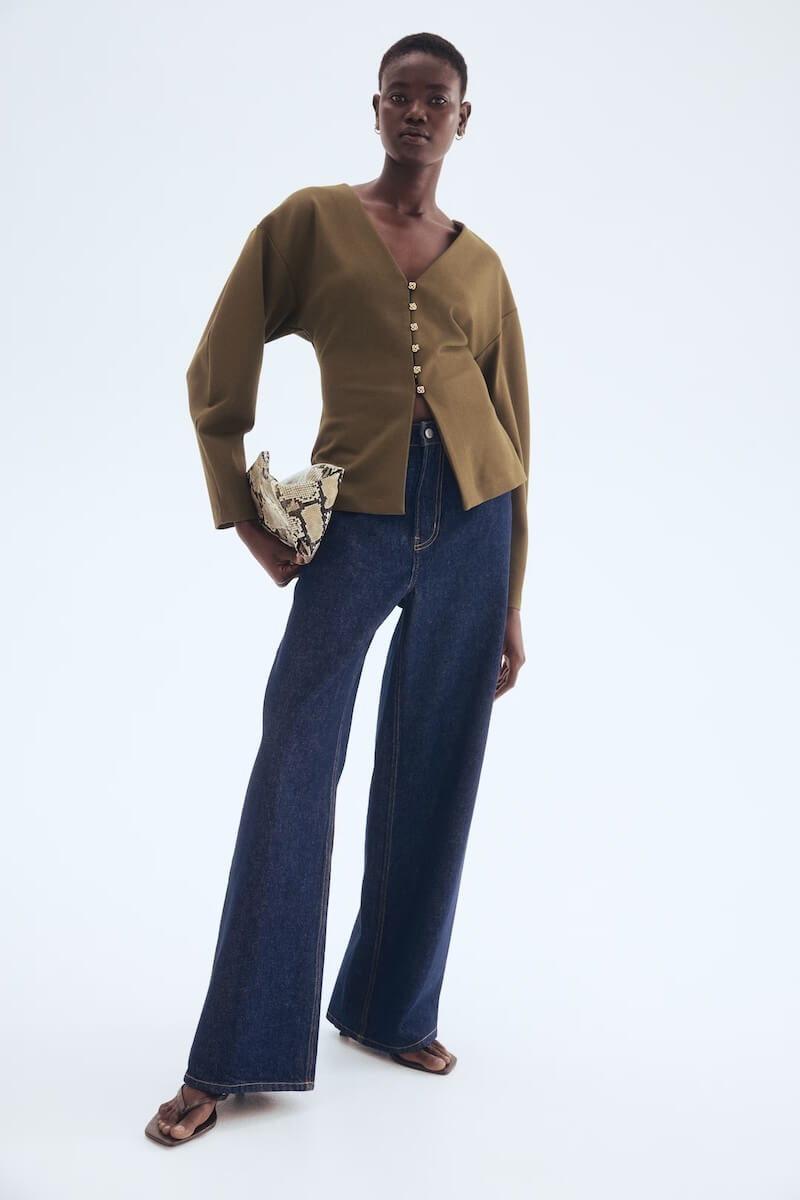 Fall 2025 Fashion: A Stylist's Lookbook of This Season's Trends - 11 A person stands wearing a loose olive green button-up top, wide-leg dark blue jeans, sandals, and holding a snakeskin-patterned clutch against a plain background—an effortless nod to fall 2025 fashion.