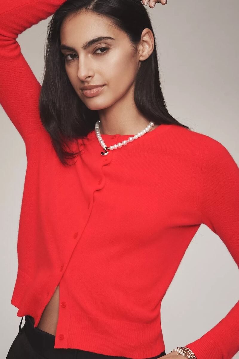 Fall 2025 Fashion: A Stylist's Lookbook of This Season's Trends - 59 A woman wearing a bright red cardigan and a pearl necklace, inspired by fall 2025 fashion, poses with one arm raised against a neutral background.