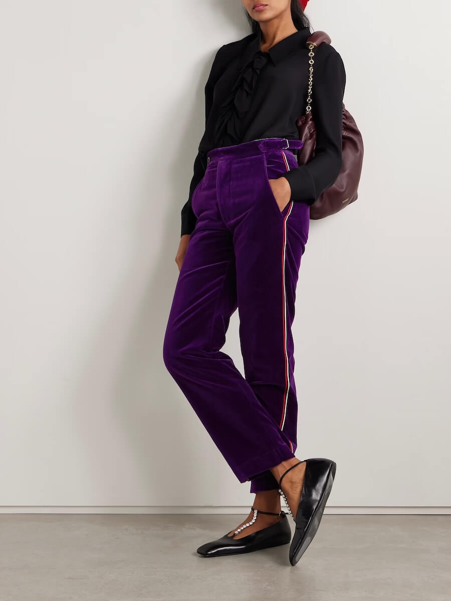Fall 2025 Fashion: A Stylist's Lookbook of This Season's Trends - 57 Person wearing a black blouse, purple velvet pants with side trim, black pointed-toe shoes, and carrying a burgundy shoulder bag—an effortless nod to Fall 2025 fashion trends—standing against a plain wall.