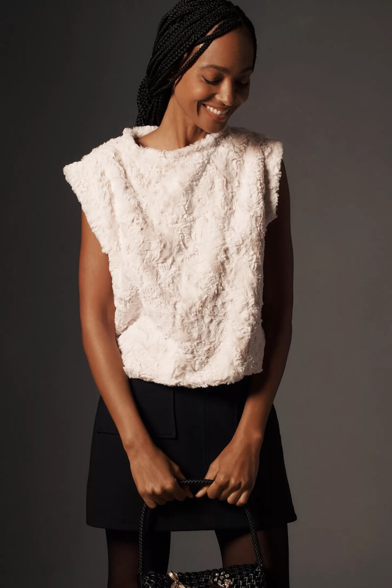 Fall 2025 Fashion: A Stylist's Lookbook of This Season's Trends - 18 Woman standing against a dark background, wearing a sleeveless textured white top and a black skirt, holding a black handbag, and smiling while looking down—an elegant look inspired by fall 2025 fashion trends.
