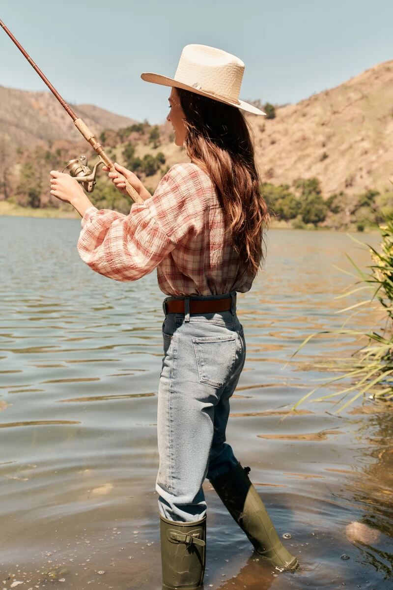 Fall 2025 Fashion: A Stylist's Lookbook of This Season's Trends - 62 A person wearing a wide-brimmed hat, plaid shirt, jeans, and rubber boots—channeling Fall 2025 Fashion Trends—stands in shallow water by a lake, fishing with a rod.
