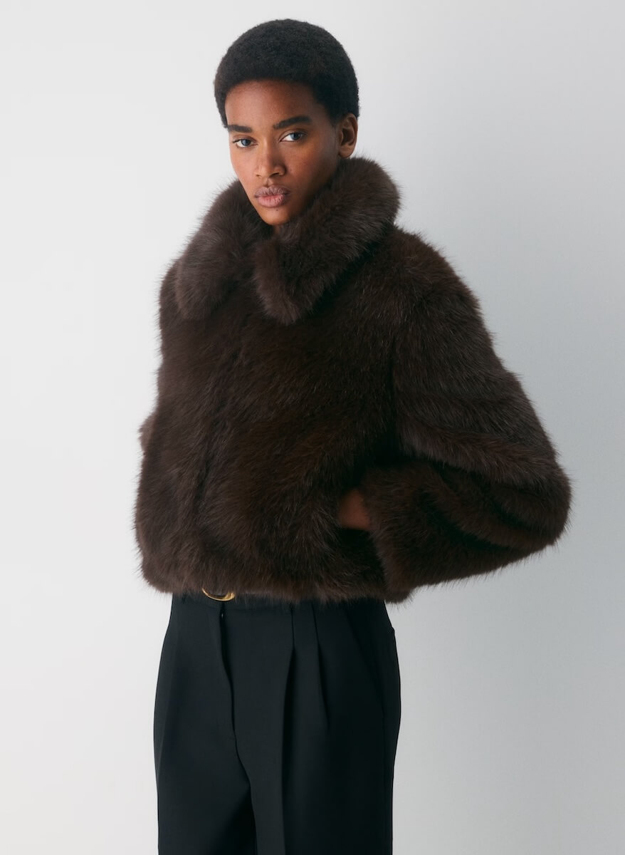 Fall 2025 Fashion: A Stylist's Lookbook of This Season's Trends - 26 A person stands against a plain background wearing a dark brown fur coat and black trousers, embodying Fall Fashion trends for 2025 with hands in their pockets and a neutral expression.