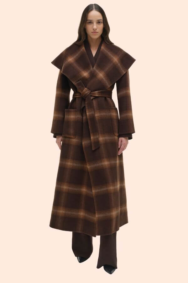 Fall 2025 Fashion: A Stylist's Lookbook of This Season's Trends - 77 A woman stands facing forward wearing a brown plaid belted coat with wide lapels, brown pants, and black shoes—a chic look that captures Fall 2025 fashion trends against a plain light background.