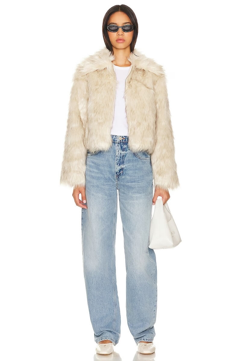 Fall 2025 Fashion: A Stylist's Lookbook of This Season's Trends - 29 A woman wearing a light faux fur jacket, white t-shirt, loose blue jeans, white sneakers, sunglasses, and holding a white handbag stands against a plain background—perfectly capturing effortless fall 2025 fashion.