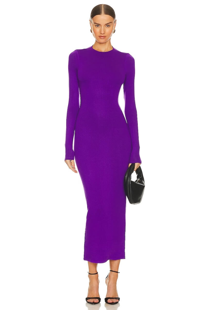 Fall 2025 Fashion: A Stylist's Lookbook of This Season's Trends - 56 A woman wears a long-sleeve, form-fitting purple dress with a crew neckline—an elegant look inspired by fall 2025 fashion—paired with a black handbag and high-heeled sandals, standing against a white background.