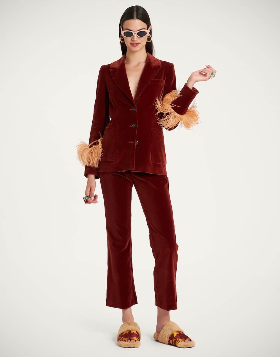 Fall 2025 Fashion: A Stylist's Lookbook of This Season's Trends - 53 A woman wearing a burgundy velvet pantsuit with feathered cuffs, patterned slippers, sunglasses, and jewelry stands against a plain white background, perfectly capturing Fall 2025 fashion trends.