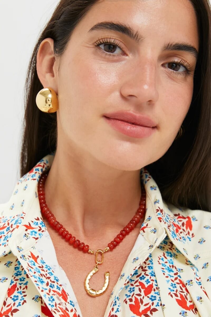 Fall 2025 Fashion: A Stylist's Lookbook of This Season's Trends - 40 Woman wearing a cream blouse with a floral pattern, large round gold earrings, and a red beaded necklace with a gold horseshoe pendant—perfect for showcasing fall 2025 fashion trends.