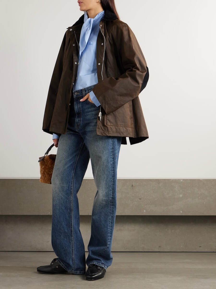 Fall 2025 Fashion: A Stylist's Lookbook of This Season's Trends - 74 Embracing Fall 2025 fashion trends, a person wears a brown jacket, blue shirt, wide-leg jeans, and black shoes while holding a small brown textured bag, standing indoors against a plain background.