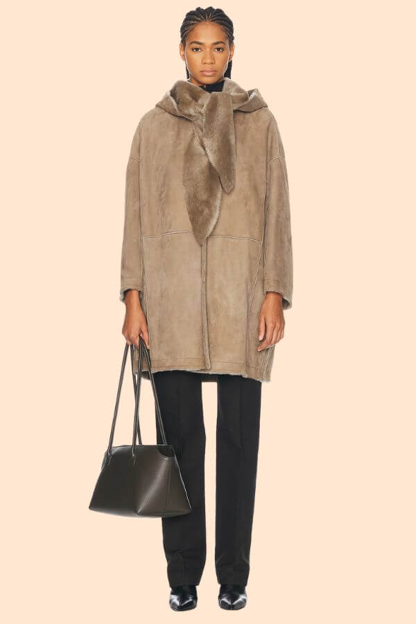 Fall 2025 Fashion: A Stylist's Lookbook of This Season's Trends - 32 A woman wearing a brown oversized shearling coat, black pants, and black shoes, holding a black handbag, stands against a beige background—an effortless nod to Fall 2025 fashion trends.