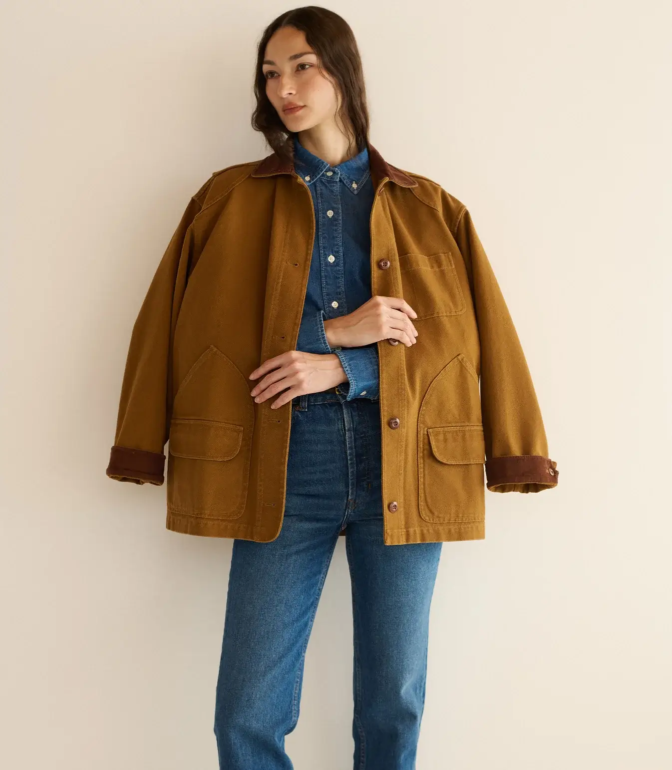 Fall 2025 Fashion: A Stylist's Lookbook of This Season's Trends - 65 A woman stands against a beige wall wearing a brown jacket with large pockets over a denim shirt and blue jeans, channeling effortless fall 2025 fashion.
