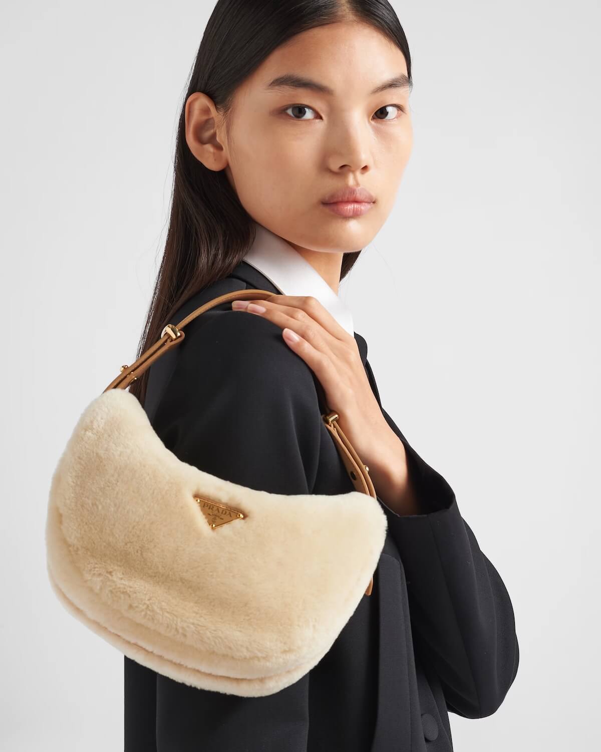 Fall 2025 Fashion: A Stylist's Lookbook of This Season's Trends - 36 A person with long dark hair wearing a black blazer holds a cream-colored fuzzy handbag with a triangular logo on the front, showcasing one of the top Fashion Trends for Fall 2025.