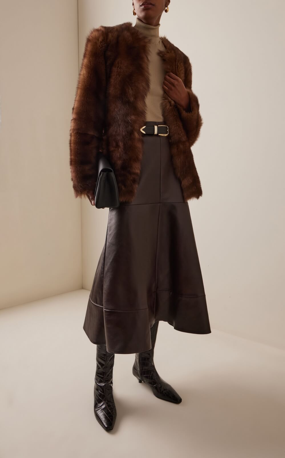 Fall 2025 Fashion: A Stylist's Lookbook of This Season's Trends - 23 Showcasing Fall 2025 fashion trends, this look features a brown fur coat, beige turtleneck, brown leather skirt, black belt and boots, with a chic black clutch—perfectly styled against a plain light-colored wall.