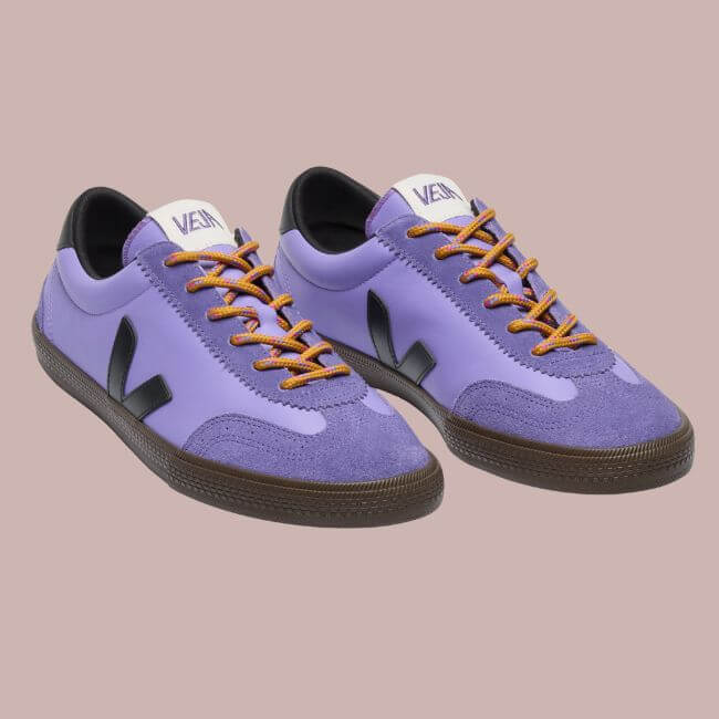 Fall 2025 Fashion: A Stylist's Lookbook of This Season's Trends - 48 A pair of purple Veja sneakers with black soles, black side logos, orange laces, and white tongue labels on a plain light mauve background—perfect for embracing Fall 2025 fashion trends.