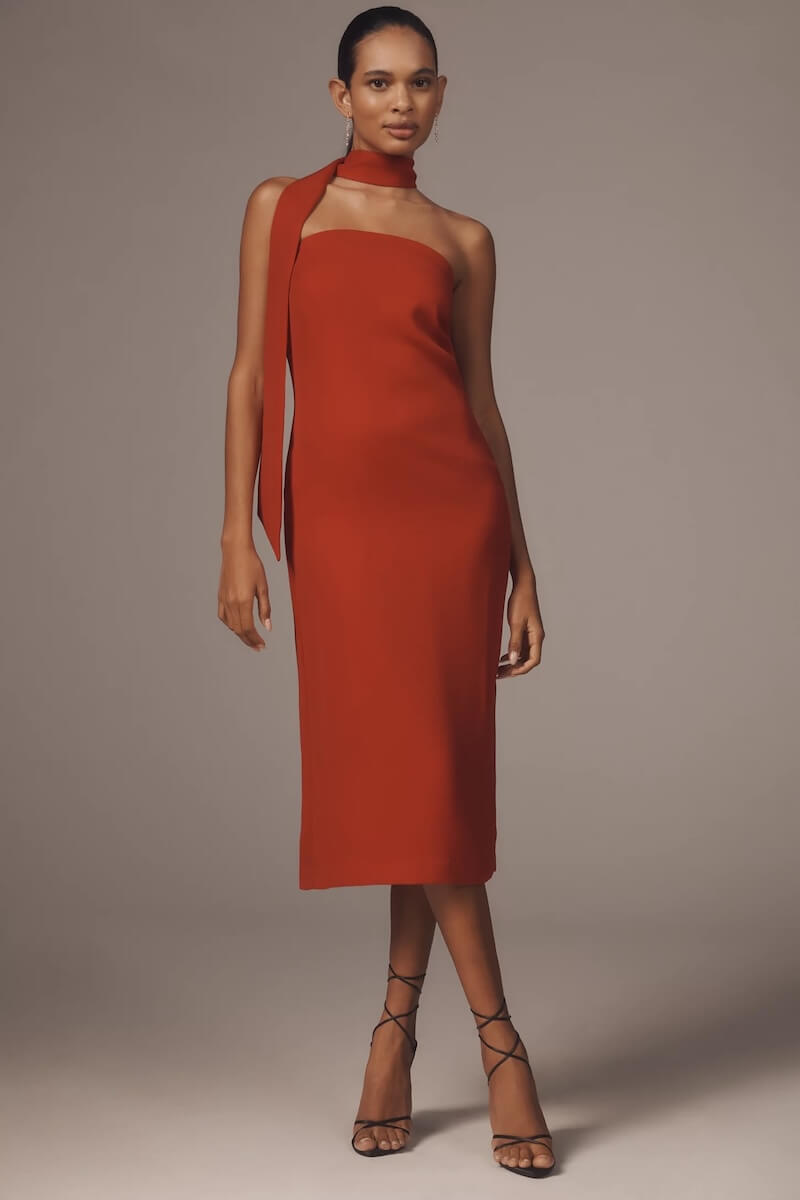 Fall 2025 Fashion: A Stylist's Lookbook of This Season's Trends - 49 A woman stands wearing a sleeveless, knee-length red dress with an attached necktie detail and black strappy high-heeled sandals, embodying chic fall 2025 fashion against a plain background.